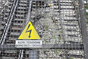 Yellow warning sign for high voltage contact wires above the rails, in the background the train station tracks. Warning icon