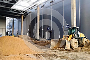 A yellow wheel loader maneuvers a heap of soil on an industrial construction site. Steel beams support the skeletal