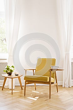 Yellow wooden armchair next to table in white living room interior with drapes. Real photo