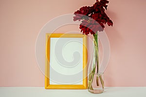 Yellow wooden photo frame mockup, red cynicism in glass vase in front of pale pink pastel background.