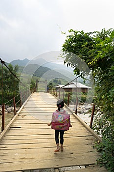 Yen Bai, Vietnam - Sep 18, 2016: Vietnamese Hmong ethnic minority girl walking home on old small chain wooden bridge from school w