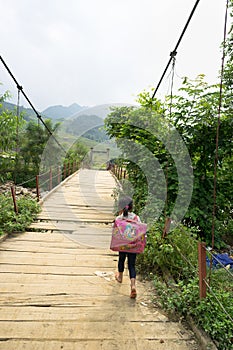 Yen Bai, Vietnam - Sep 18, 2016: Vietnamese Hmong ethnic minority girl walking home on old small chain wooden bridge from school w