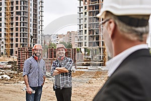 Yes, boss Professional team of two young cheerful builders are standing at construction site in helmets and smiling to