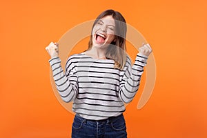 Yes I did it! Portrait of joyous winner, young woman with brown hair in long sleeve striped shirt. indoor studio shot isolated on