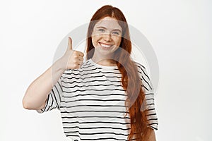 Yes. Smiling redhead female model showing thumbs up. Girl with red long hairstyle and freckles likes smth good, give