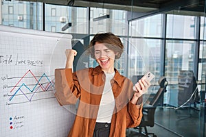 Yes, success. Happy young sales manager, woman celebrating victory, standing near office, posing near diagram, holding