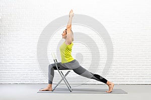 Yoga with a chair. Fit caucasian woman practice warrior pose using props in loft white studio indoor.