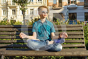 Yoga in city, teenage boy sits in lotus pose on bench in city park. Relax, rest, meditation