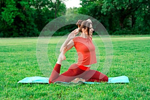 Yoga class in nature. Stretching lessons on green grass in the park. A girl is doing yoga in a pigeon pose.