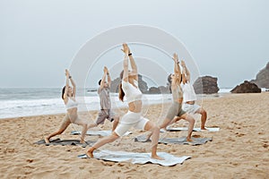 Yoga class on ocean shore. Group of young people doing warrior pose, meditating and exercising on the beach