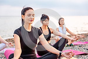 Yoga class at sea coast. Group of Caucasian women meditating on sports mats at pebble beach. Outdoor training and