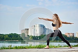 Yoga female standing in a warrior pose, Virabhadrasana, near river in background of urban buildings with copy space