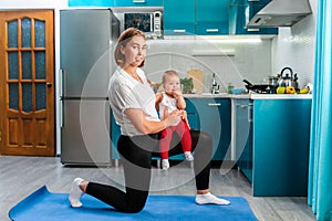 Yoga at home. A smiling young mother holding a baby in her hands and posing on a mat. Kitchen on the background. The concept of