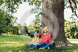 Yoga and meditation concept. Beautiful young woman in knitted clothes and small child keep eyes closed, meditate outdoors in green