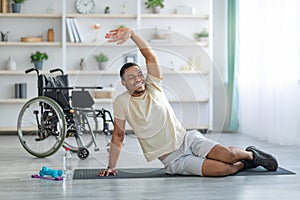 Yoga for paraplegic patients. Disabled black guy stretching on sports mat at home, wheelchair standing nearby