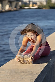 Yoga runner woman in pink legging sitting on embankment, stretching muscles in seated forward bend position. Hamstring stretch