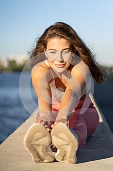 Yoga runner woman in pink legging sitting on embankment, stretching muscles in seated forward bend position. Hamstring stretch