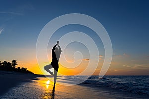 Yoga vrikshasana tree pose by woman in silhouette on the beach with sunset sky background. Free space for text.