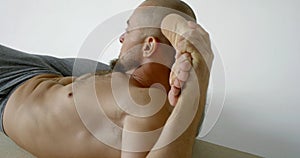 Yogi man is holding leg behind head, lying on floor, stretching muscles
