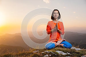 Yogini in the lotus position in full face sits on a cliff against the backdrop of the sunset sky with mountains, prays