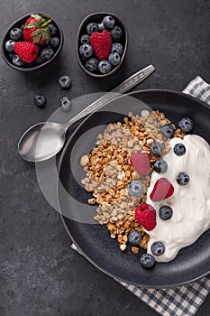 Yogurt with baked granola and berries in black ceramic plate on dark stone background. Healthy breakfast. Vertical photo