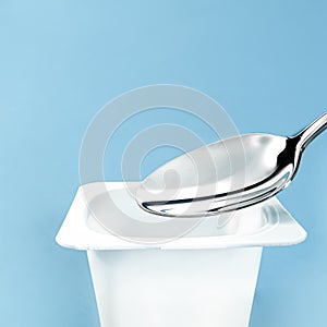 Yogurt cup and silver spoon on blue background, white plastic container, fresh dairy product for healthy diet and