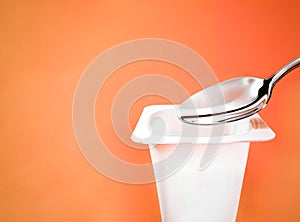Yogurt cup and silver spoon on orange background, white plastic container with yoghurt cream, fresh dairy product for