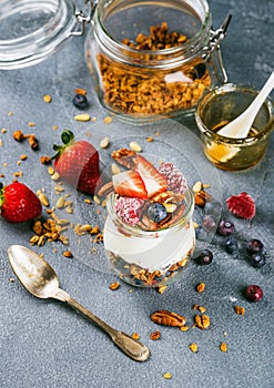 Yogurt oat granola with fresh berries, nuts, honey and mint leaves in glass jar on grey concrete textured backdrop