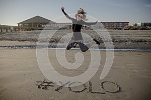 YOLO hashtag written in the sand on the beach and an adult female jumping.