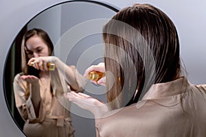 Yong Woman Brunette Applying Essential Oil Spray On Her Hair Standing Near Mirror In Bathroom. Split Ends Repair Treatment.