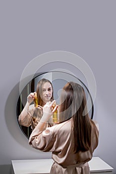 Yong Woman Brunette Applying Essential Oil Spray On Her Hair Standing Near Mirror In Bathroom. Split Ends Repair Treatment.