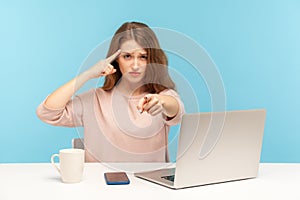 You are idiot! Woman with sarcastic look sitting at workplace and making stupid gesture, pointing to camera