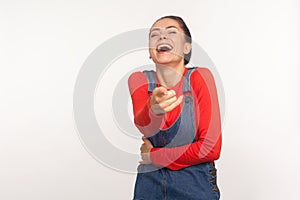 You look ludicrous! Portrait of girl in denim overalls bursting into laughing and pointing finger to camera, teasing photo