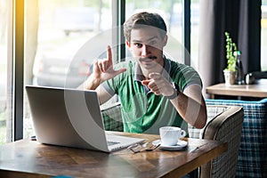 You are loser. Young serious businessman in green t-shirt sitting, pointing and showing loser gesture and looking at camera