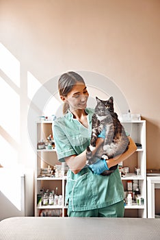 Are you scared Pleasant young female vet holding a big black cat and smiling while looking at him while standing at the