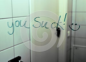 `You suck!` written on a bathroom mirror. A happy smiley ends the sentence