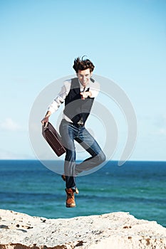 You too could have this freedom. Handsome young man holding a briefcase jumping in the air with the ocean in the