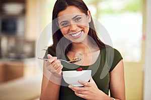 You are what you eat. Portrait of a happy young woman eating a bowl of muesli while standing in her kitchen.