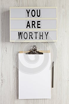 `You are worthy` on a lightbox, clipboard with blank sheet of paper on a white wooden background, top view. Flat lay, overhead,