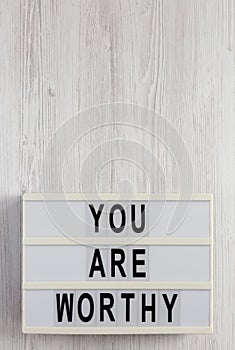 `You are worthy` on a lightbox on a white wooden background, top view. Flat lay, overhead, from above. Copy space
