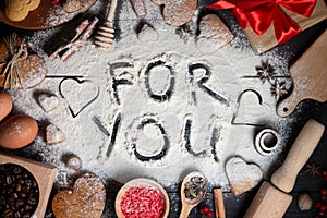 For You written on flour. Gingerbread heart shaped cookies, spices, baking supplies on black wood background
