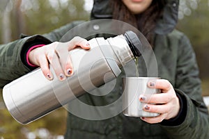 Youg beautifull women pouring beverage from termos to cup in a cold raini day. Healthy living and hiking