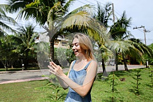 Youg caucasian girl talking by sartphone near palm trees and road, wearing jeans sundress.