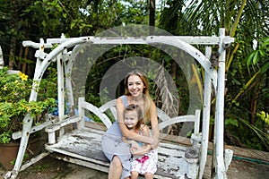 Youg mother with daughter riding on swing in exotic garden, palms in background.