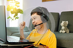 Youn little cute Asian 10s boy wearing headphones sitting in home living room using laptop notebook computer for online distance
