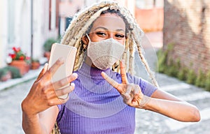 Yound african woman with blond dreadlocks doing video call with smart mobile phone wearing face protective mask - Technology and