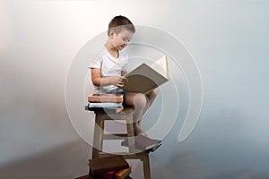 Yound kid reading interesting book at the wooden chair near white wall. Concept of reading an interesting and exciting book everyw