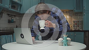 Young 20s man in a hurry looking at laptop and making last sip of tea and going away from blue kitchen. Male student at