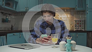 Young 20s man surfing online using mobile phone and smiling sitting at the dining table on blue kitchen. Male with