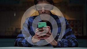 Young 20s man using mobile phone in hands sitting in dark. Man`s hands with cellphone in focus.